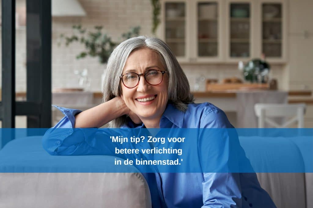 Vrouw, 50+ binnen zittend op de bank en lachend in de camera. Tekst: 'Mijn tip? Zorg voor een betere verlichting in de binnenstad.'